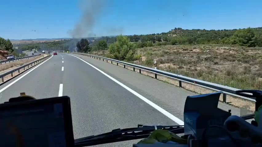 The video shows a view from inside a vehicle traveling on a highway. Smoke is visible rising from vegetation to the right of the highway in the distance. Several individuals wearing high-visibility yellow vests are standing on the shoulder of the highway near the source of the smoke. The terrain is dry and hilly, with scattered trees and brush. The sky is clear and blue. The highway appears to be a multi-lane road with a metal guardrail separating the lanes from the shoulder.