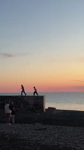 Two children are running on a concrete barrier at a beach during sunset. There is graffiti on the barrier. The sky is a gradient of blue, orange, and pink. A small group of adults is standing on the beach near a stroller, observing the children. The beach is covered in small dark rocks. The sea stretches to the horizon.
