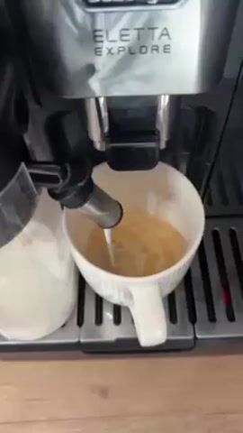A DeLonghi Eletta Explore coffee machine is shown dispensing what appears to be steamed milk into a white mug of coffee. The mug is on the machine's drip tray, and the milk is being dispensed from an attachment on the left side of the machine. The background is a light-colored surface, possibly a countertop. The scene is well-lit.