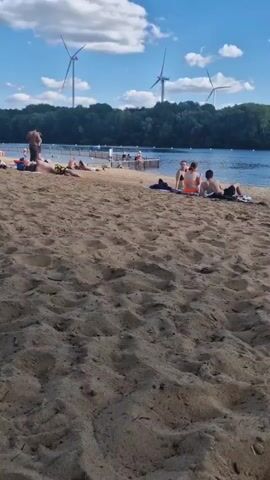 The video opens on a sandy beach with several people relaxing. In the background, a body of water is visible with a wooden barrier extending into it, where more people are gathered. Some individuals are sitting on the barrier, with their legs in the water. Further in the distance, a dense line of trees forms the horizon, and several wind turbines are visible against a partly cloudy sky. The camera pans slightly to the right, focusing on groups of people on the beach and by the water. Some are sunbathing, while others are seated by the water. The overall scene depicts a leisurely day at a beach or lakefront.
