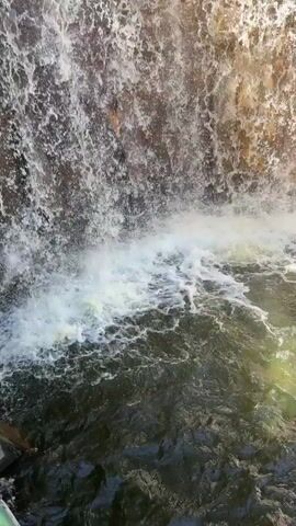 The video shows a close-up view of a waterfall. Water cascades down a rough, textured rock face into a pool below. The water is white and frothy where it impacts the pool, which is dark and slightly reflective, with hints of green from sunlight. The rock face appears to be damp and covered in varying tones of gray and brown.