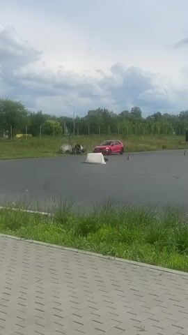 A red Volkswagen Golf GTI is seen performing a drift on a paved track. The car is angled sideways as it navigates a curve, with its rear wheels losing traction. In the background, a white rectangular obstacle, several orange traffic cones, and green foliage are visible. The sky is partly cloudy. The camera is positioned at a low angle, looking towards the track from the side, with some grassy area and paving stones in the foreground. The car appears to be the primary subject, engaged in a dynamic driving maneuver on an open area.