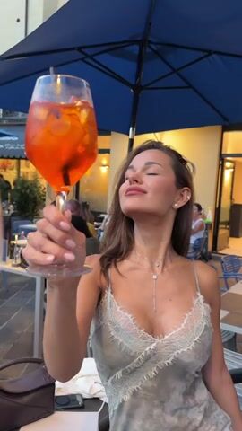 The video begins with a close-up of a woman holding a glass of an orange cocktail with ice and a slice of orange. She is wearing a light-colored, sleeveless dress with lace trim. She tilts her head back and smiles, then brings the glass closer to her face. The background shows an outdoor dining area with tables, chairs, and umbrellas. Other patrons are visible in the background, some sitting at tables and others walking. The lighting suggests it is evening. The video then pans out to show more of the outdoor dining area, with more tables and people. The focus shifts to a group of people sitting at a table in the distance. The video ends with a slightly blurred shot of the outdoor dining area and the people in it.