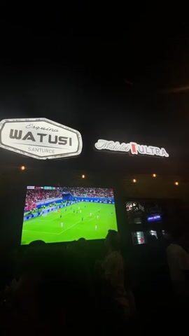 The video shows a nighttime scene inside what appears to be a bar or restaurant. Above a large screen, two illuminated signs are visible: "Esquina WATUSI SANTURCE" on the left and "Michelob ULTRA" on the right. The large screen displays a soccer match with players in white and red uniforms on a green field. Below the screen, the silhouettes of several people are visible, suggesting they are watching the game. The overall atmosphere is dark, with some lighting provided by the screen and the signs.