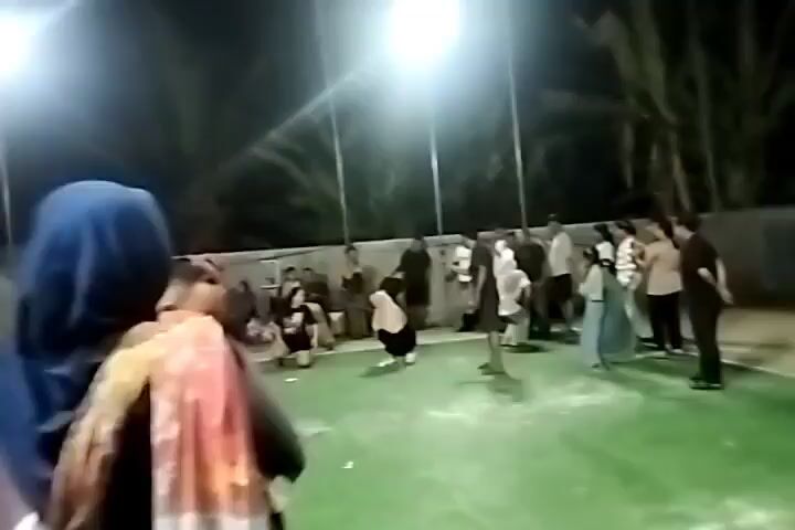 The video shows a group of people gathered outdoors at night on what appears to be a sports court. The court has a red and green surface, with white lines marking boundaries. Several bright floodlights illuminate the scene. In the foreground, from a slightly elevated perspective, a person wearing a blue hijab and a colorful scarf is visible from behind. They are looking towards the court where various individuals are engaged in activity. In the middle ground, a group of people are interacting. Some are standing, while others are seated on the ground. A person in a red shirt is walking across the court. A woman in a white hijab is kneeling. Another person in a white shirt and dark pants is also on the court. In the background, more people are gathered, some sitting and watching, others standing and talking. Palm trees and a fence are visible behind the court. The overall atmosphere suggests a social gathering or recreational activity taking place in an outdoor court area at night.