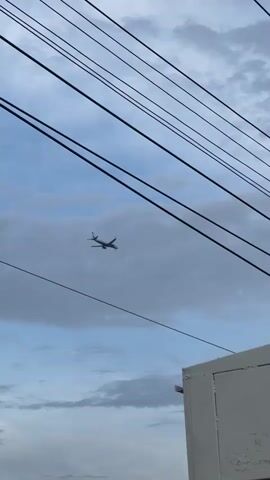 The video shows an airplane descending in the sky. The plane is framed by overhead power lines and the roof of a building labeled "TRAFFIC OFF". The sky is cloudy.