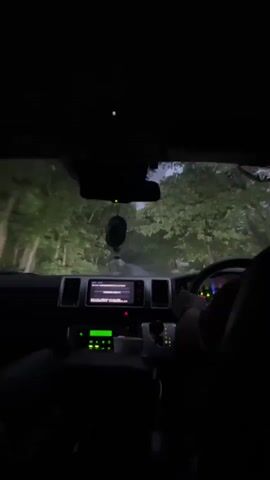 The video shows the interior of a vehicle driving on a road surrounded by trees at night. The windshield wipers are in motion, indicating it might be raining. The dashboard lights are on, and the navigation screen is visible. The driver's hands are on the steering wheel. The perspective is from inside the vehicle, looking forward.