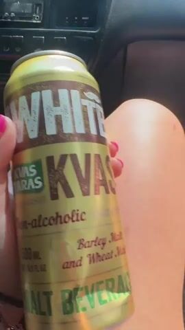 The video begins with a close-up of a person's hand holding a can of "WHITE KVAS Non-alcoholic Barley Malt and Wheat Malt" beverage. The hand has bright pink fingernails. The scene then shifts to a view from inside a car, looking out through the windshield. The car appears to be moving along a road surrounded by trees and foliage. In the distance, a tall communication tower is visible against a clear blue sky. The interior of the car shows the dashboard, including the radio and air vents. The perspective suggests the video is a first-person point of view from a passenger.