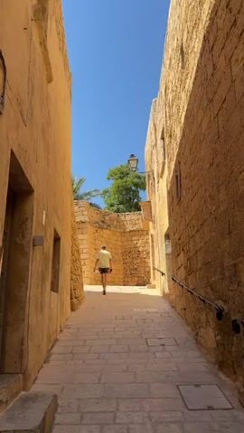 A person wearing a yellow and white patterned shirt and shorts walks away from the camera down a narrow, sunlit stone alleyway. The walls of the alley are constructed from large, rough-hewn yellow stone blocks, creating a textured, ancient appearance. The ground is paved with light-colored cobblestones. In the background, a large green tree is visible above the stone wall, and the sky is a clear, bright blue. As the video progresses, another person wearing a white long-sleeved shirt and white shorts, and a tan bucket hat, walks into the frame from the right. A person wearing a white t-shirt with a cloud pattern, sunglasses, and a white baseball cap walks behind the person in the bucket hat, looking at them. They are walking in the same direction as the first person. The two people walking together are seen from behind, moving down the alleyway.