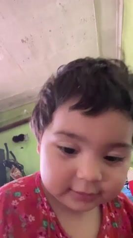 The video shows a close-up of a young child's face. The child has dark hair and is wearing a red shirt with a floral pattern. The child's expression changes from neutral to surprised, then to a smile. The background appears to be an indoor room with green walls and a white ceiling. There are some objects visible in the background, including what looks like a mirror, some bags, and a lamp. The lighting is somewhat dim.