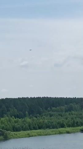 A video clip shows a landscape from above. In the foreground is a body of water. Beyond the water is a line of trees, followed by more trees and a cloudy sky. There are several objects in the sky that appear to be small aircraft or drones. The video is 4 seconds long.