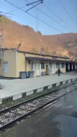 A train is pulling away from a train station. The station is a low, rectangular, light yellow building with a dark roof. A Bulgarian flag is displayed on the building. A man in dark clothing is standing on the platform. Behind the station is a low, wooded mountain. The sky is blue. The video is shot from inside the train.