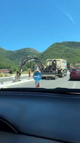 The video shows a view from inside a vehicle, looking out at a road construction site. In the foreground, the dashboard and windshield of the vehicle are visible. In the middle ground, a Volvo excavator is positioned on the side of the road, with a large dump truck behind it. A blue and white traffic sign with a white arrow pointing to the right is also visible. The road appears to be under construction, with barriers and unfinished sections. In the background, green hills and houses can be seen under a clear blue sky. Several other vehicles are also on the road, including a red car and a black car. The excavator operator is visible in the cab of the machine.