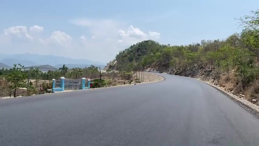 The video shows a view from a black paved road curving to the right. On the left side of the road is a partially constructed area with a blue and white fence. Beyond the fence is a view of a body of water, palm trees, and mountains in the distance under a blue sky with white clouds. The right side of the road is bordered by a hillside with trees and shrubs. A sign reading "HO SONG LUY" is visible in the distance beyond the fence. Power lines are visible stretching across the landscape.