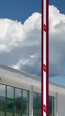 The video features three brief upward shots of a white building on a clear, sunny day. The sky is a vibrant blue, adorned with large, puffy white cumulus clouds.

The first shot focuses on the corner of the white building, showcasing its flat roofline and several green-framed windows. Two security cameras are visible on the exterior wall. The second shot introduces a tall, grey metal pole in the foreground, positioned centrally and partially obscuring the building and clouds behind it. The final shot captures the top part of the white building, revealing a prominent red sign with the text "VAVILOV" (partially visible as "VAVILO") emblazoned on its facade.

The setting is outdoors, likely a commercial or public building given the sign. The weather is bright and clear, indicative of daytime. The location, as per context, is Primorskoe, Abkhazia. There are no people or specific activities depicted.