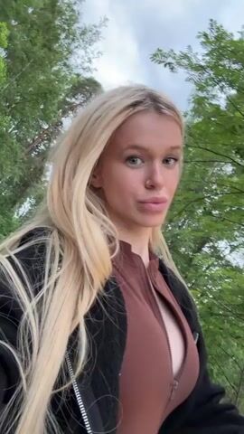 A young woman with long blonde hair, wearing a brown zippered bodysuit and a black jacket, is walking outdoors. She is looking at the camera and smiling. The background consists of trees and foliage. The camera appears to be moving along with her.