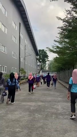 A group of people, mostly women wearing hijabs and casual clothing, walk away from the camera down a paved walkway. The walkway is bordered on the left by a long, gray, multi-story building with numerous windows, and on the right by a fence and trees. Some individuals carry bags or packages. The sky is overcast.