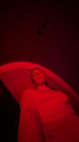 A low-angle shot shows a young woman posing in a room lit with a strong red light. She is wearing a light-colored top, possibly white or cream, and she flicks her hair. The background is dark and undefined, with hints of walls or furniture. The red light creates a dramatic and somewhat abstract effect, obscuring details and focusing attention on her face and movements. Music plays in the background.