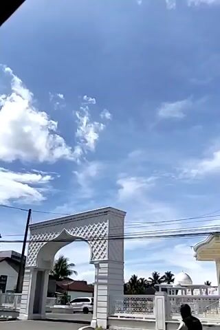The video shows a white mosque under a bright blue sky dotted with clouds. The mosque features multiple black domes and arched windows. A white archway stands on the left side of the frame, and power lines run across the view. There are also some trees and a car visible in the background.