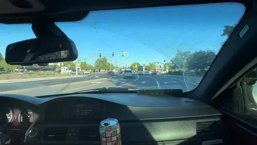 The video shows the interior of a car driving on a sunny day. The camera is mounted inside the car, facing forward through the windshield. The windshield has some streaks and smudges on it. In the foreground, the dashboard is visible, including the steering wheel and a part of the instrument cluster. A can of soda with a colorful design is perched on the center console. The view through the windshield shows a road with multiple lanes and other cars. Traffic lights are visible at an intersection ahead. Trees and buildings line the sides of the road. The sky is clear and blue. The driver's left hand is visible on the steering wheel. A rearview mirror is also visible, reflecting the interior of the car. The overall scene depicts a typical driving situation on a bright day.