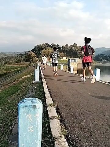 The video shows an outdoor scene, likely in Jambeyan, Indonesia, depicting several young people walking or jogging along a paved path. The path appears to be an embankment or a road running alongside a body of water, possibly a lake or reservoir, visible on the right side of the frame.

To the left of the path is a grassy bank. Blue and white concrete posts line both sides of the path, acting as markers or part of a railing system. In the background, there's a hill covered with dense trees. Further in the distance, more hills or mountains are visible under a clear, bright sky, suggesting daytime, possibly late afternoon.

One person in a white shirt leads, followed by another wearing a black and white jersey with the number "28" on the back. A third person, wearing a red shirt and a small backpack, is jogging behind them. A structure with blue railings is visible near the water further down the path. The overall atmosphere is calm and pleasant, indicating a leisurely or exercise activity.