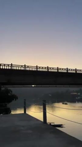 The video shows a bridge silhouetted against a twilight sky over a body of water. The sky fades from blue at the top to a pale yellow near the horizon. The bridge has a distinctive railing with repeating arched details. Below the bridge, the water reflects the golden light of the sky. A concrete platform with wooden posts and rope sits in the foreground on the left. Trees and buildings are visible on the opposite shore, and a couple of boats are on the water.