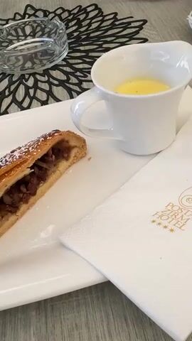 The video shows a close-up of a dessert on a white rectangular plate. The dessert is a slice of apple strudel, sprinkled with powdered sugar, next to a small white pitcher filled with yellow custard sauce. To the right of the plate is a white napkin with a gold logo that reads "POSTA ZIRM HOTEL" and four gold stars. The table surface is a light-colored wood grain. In the background, a black and white patterned placemat and a glass ashtray are visible. The camera then pans to the right to reveal a black bowl filled with chocolate ice cream or mousse, topped with whipped cream and chocolate drizzle. A woman with blonde hair, wearing a black tank top and a light-colored skirt, is seated at the table, holding a smartphone and looking at it. She is wearing a gold necklace, large gold hoop earrings, and has long, manicured nails. In the far background, through glass doors, a store window display with clothing and sunglasses can be seen. The scene appears to be an outdoor seating area of a cafe or restaurant.