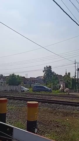 The video captures a train moving on a railway track through a rural area on a sunny day. The view is slightly low, focusing on the train's passage. The train, identified as a KAI (Indonesian Railway Company) train, is predominantly white with horizontal blue and orange stripes along the carriages. The locomotive has the code CC2010405. The train is moving from left to right in the frame. The railway track is visible in the foreground with gravel and some sparse vegetation. Wooden posts with yellow and red tops are placed alongside the track. In the background, there are houses, trees, and overhead power lines, creating a typical countryside scene. The sky is clear and bright.