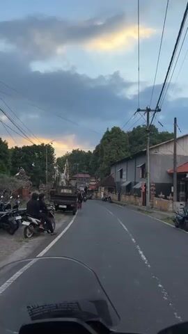 The video shows a perspective from the handlebars of a motorcycle as it travels down a paved road. The road is bordered by trees and buildings, with power lines running overhead. Several motorcycles are parked along the side of the road, and some are seen in motion. There are also a few cars and a pickup truck visible. The sky is cloudy with a soft glow of sunset visible in the distance. On the left side of the road, there is a large statue and a decorative structure, possibly related to a cultural event. The overall impression is that of a rural or suburban street scene in a tropical climate.