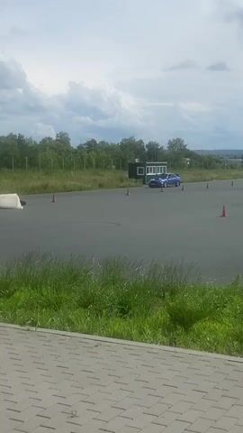 A blue Subaru Impreza WRX STI with a large rear spoiler is drifting around a paved track. Orange traffic cones are placed around the track, some of which the car passes close to. A white concrete barrier is also visible on the track. In the background, there are trees and a small building. The sky is cloudy. The camera is positioned from a slightly elevated viewpoint, looking down on the track.