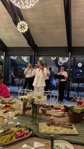 A full-frame, medium shot captures a band performing in a dimly lit room, likely a restaurant or event hall. The camera is positioned at table height, focusing on the food spread in the foreground and the performers in the background. A woman in a white suit stands center, singing into a microphone with her right hand raised. To her left, a man in a white shirt plays a trumpet, and to her right, another man in a white shirt plays a saxophone. A fourth musician, in a dark shirt and black pants, is partially visible playing a saxophone behind the singer. The room features a wooden ceiling with hanging spherical light fixtures. Tables in the foreground are laden with various dishes, including fruits, vegetables, and what appears to be cheese. The overall atmosphere suggests a lively indoor event.