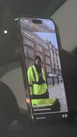 A smartphone, resembling an iPhone 14 Pro/Pro Max, is positioned vertically, likely mounted in a car with parts of the dark interior visible in the foreground and background. The time displayed on the phone's status bar is 22:46.

The phone is playing a video on a social media platform. The video shows a man standing outdoors on a sidewalk, wearing a black beanie, a black neck gaiter or scarf, and a bright yellow/lime green high-visibility jacket with reflective stripes. Behind him, there are multi-story buildings with classic architecture, consistent with urban European settings, and other people walking in the distance.

Visible text overlays on the video include the user handle "Auditing Britain" with a "Follow" button, and a caption or title that reads "British Broadcasting Garbage". Engagement metrics are also visible, showing 2.7K likes and 318 comments.

The audio accompanying the video features the man speaking about people stopping payment for a "license," and he states, "which I am one of them." A green light is faintly visible in the upper left of the overall scene, possibly an indicator light within the car.