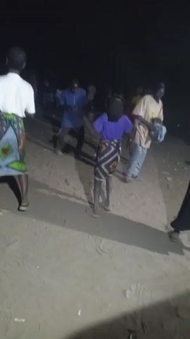 In a dimly lit outdoor setting, a group of people are gathered, seemingly for a social event. Several individuals are actively dancing. The person closest to the camera, a slender individual wearing a purple shirt and a patterned wrap skirt, is dancing facing away from the camera. To their left, a man in a blue shirt is also dancing. Further in the background, another woman in a red shirt dances alongside the first person. The ground appears to be dirt or packed sand. Other people are visible in the background, though their details are less clear due to the low light and distance.
