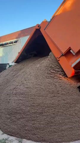 A dump truck is shown with its bed tilted upwards, unloading a large pile of sand. The sand is a light brown color and has a granular texture. The truck is orange and black, and its tires are visible. The background shows a building with scaffolding and a clear blue sky. The scene appears to be outdoors, possibly at a construction site or a material yard. The action captured is the continuous pouring of sand from the truck's bed. There are no people visible in the frame. The lighting suggests it is daytime, likely late afternoon or early morning due to the long shadows and the angle of the sun.