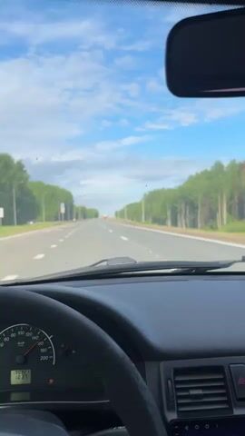 The video shows a first-person perspective from inside a car driving on a highway in Beloborodovo, Russia. The car's dashboard, steering wheel, and part of the rearview mirror are visible in the foreground. The car appears to be a Lada, indicated by the instrument cluster design. The speedometer reads approximately 80 km/h, and the odometer shows 19092.1 km. A digital display on the right of the dashboard indicates "Bluetooth" connectivity.

The highway is straight and wide, with two lanes in the direction of travel and a median. It is flanked by dense green trees on both sides, suggesting a scenic route. Streetlights are intermittently spaced along the road. The sky is bright blue with scattered white clouds, indicating clear daytime weather. In the distance, a red car can be seen. Russian speech is audible in the background.