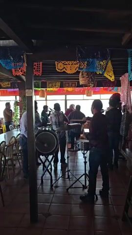 A group of musicians are playing in a restaurant with a view of the ocean. The musicians are playing drums and brass instruments. The restaurant is decorated with papel picado banners. There are tables and chairs in the foreground. The scene is well-lit by natural light from the windows.