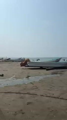 The video shows a beach scene with a sandy shore, the ocean, and a clear sky. Several small, white boats are beached on the sand. Some are closer to the camera and others are further away.  In the ocean, there are several people swimming or standing in the water. A few birds are visible flying in the sky. There are small pools of water on the sand near the boats.