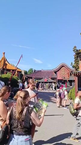 The video shows a sunny day with a bright blue sky. Several people are walking along a paved path. In the background, there are buildings with traditional Chinese architecture, adorned with red lanterns and intricate carvings. One building has a distinctive curved roof. To the left, a tall red pole with a flag or banner is visible. A wooden structure resembling a tower stands near some greenery. People are dressed in casual summer clothing. Some are looking at their phones, others are talking, and some are carrying backpacks. The atmosphere appears to be that of a tourist attraction or theme park.