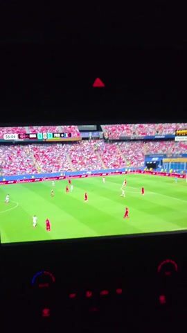 This is a video of a soccer match being displayed on a screen, likely a car's infotainment system, given the surrounding dashboard controls. The match features two teams, one in red and white uniforms, and the other in white and red uniforms. The scoreboard at the top of the screen indicates the time as 55:04 and shows the score as NOR 1 - 3 FRA. The stadium is full of spectators wearing predominantly red and white clothing, suggesting a lively and engaged audience. The field is a bright green soccer pitch, and the weather appears to be good, allowing for an outdoor event. The lighting suggests it could be daytime. The screen displays a game in progress, with players actively moving around the field, some with the ball. The overall atmosphere seems to be one of excitement and focus on the game. The visible text confirms it is a soccer match, with team abbreviations and scores.