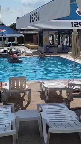 The video shows a sunny day at an outdoor swimming pool area. In the foreground, several white lounge chairs are visible, some with small tables. The pool itself is filled with blue water, and a few people are in it. One person is on an inflatable tube, and another is partially submerged. In the background, there is a covered area with a Pepsi sign, suggesting a concession stand or bar. More lounge chairs and umbrellas are scattered around the pool. Two individuals, appearing to be children, are running out of the water and onto the pool deck. The environment is clean and appears to be a public recreational space. The lighting is bright, consistent with daylight.