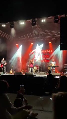 A rock band performs on an outdoor stage at night. Red and white lights illuminate the stage, with smoke effects adding to the atmosphere. The band members are on stage, with a lead singer in the center wearing a red shirt and dark pants. To the left, a guitarist in a black shirt stands near a microphone stand. Behind the singer, a drummer is visible behind a drum set. To the right, a keyboardist plays a keyboard. A large banner with text that appears to read "DIFFERENT TIMES" hangs behind the band. In the foreground, an audience is gathered, with several people silhouetted against the stage lights. Some audience members are holding up phones, presumably to record the performance. The ground in front of the stage is paved, and some stage equipment, including speakers, is visible.