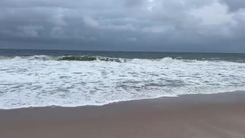 The video shows a person riding a horse on a beach. The sky is overcast with dark clouds, and the ocean has large waves crashing onto the shore. The horse is dark brown with a rider wearing a dark green shirt, grey pants, and a black cap. The horse is walking along the water's edge, with the waves coming up around its legs. The sand on the beach is a light brown color. The overall mood of the video is dramatic and somewhat moody due to the dark sky and rough sea.