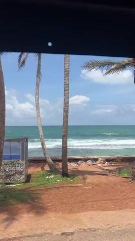 The video shows a view from inside a vehicle looking out at a tropical beach scene. In the foreground, a colorful tuk-tuk adorned with ice cream advertisements is parked on a sandy road. To the right, the ocean with gentle waves is visible under a bright, cloudy sky. Further down the beach, a small wooden kiosk sells colorful inflatable rings. A silver van is parked on the sand near the water's edge. In the distance, a few people are swimming in the ocean. The overall impression is a sunny day at a seaside tourist destination.