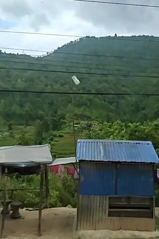 The video shows a wide shot of a hilly, forested landscape. In the foreground, there is a small, blue and brown structure that appears to be a roadside kiosk or shop, with corrugated metal at its base. Some orange clothing hangs on a clothesline to the left of the structure. In the middle ground, there are terraced fields, likely for agriculture, with some buildings visible on the hillside. The background is dominated by a steep, densely forested mountain. Power lines are visible stretching across the frame, obscuring parts of the scenery. The sky is overcast. The video appears to be filmed from a moving vehicle.