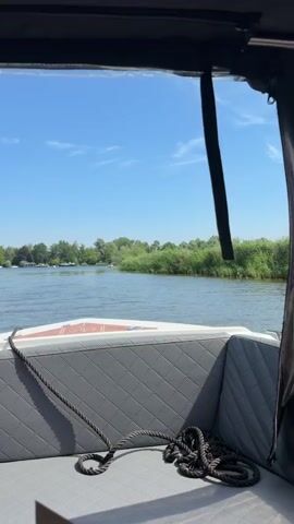 A boat's perspective shows a clear blue sky with scattered clouds. The boat has a gray quilted seat and a rope coiled on it. The boat is moving forward on a calm body of water. On the left bank of the water, there are trees. On the right bank, there is tall green grass and reeds. In the distance, on the water, there are a few other small boats visible.