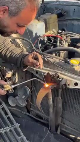 Here's a description of the video: 

A man is welding on the front of a car with the hood up. He wears a dark jacket. He holds a welding torch in one hand and uses a long, thin rod with the other. Sparks fly as he works on the metal frame in front of the car's radiator. The engine and other mechanical components are visible in the background. A detached grill sits on the ground.