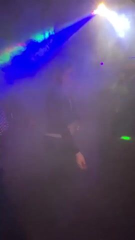 The video shows a dimly lit dance floor at a bar or club with several people dancing.  A disco ball or similar lighting fixture projects red, green, and blue light beams into the smoky room, with occasional strobe effects.  A sign reading "Doll House" is partially visible in the background.  The dancers' faces are mostly obscured by the smoke and lighting, but they are moving to the music.  One person is wearing a plaid shirt and jeans.