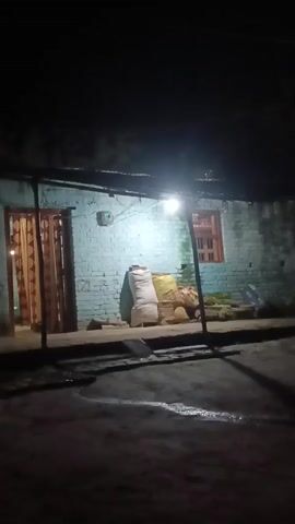 The video is a static shot of a house at night. The house has a blue brick exterior and a corrugated metal roof. A single light bulb illuminates the porch, casting shadows on the ground. There are several sacks stacked against the wall of the house. A door with a patterned curtain is visible on the left side of the porch. A window with wooden shutters is on the right side of the house. The ground in front of the house is wet and reflects the light. The overall impression is one of a simple, rural dwelling at night.
