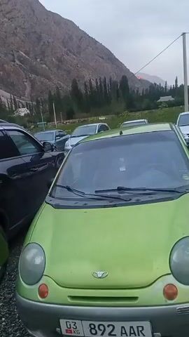 The video shows a parking lot with several cars in it. In the foreground, there is a bright green car. Behind it, other cars are parked in a row. The parking lot is gravel. In the background, there are mountains and trees. The sky is cloudy and appears to be twilight. There is a faint orange glow visible on the mountainside in the distance, possibly indicating a fire or a light source. There are no people visible in the video.