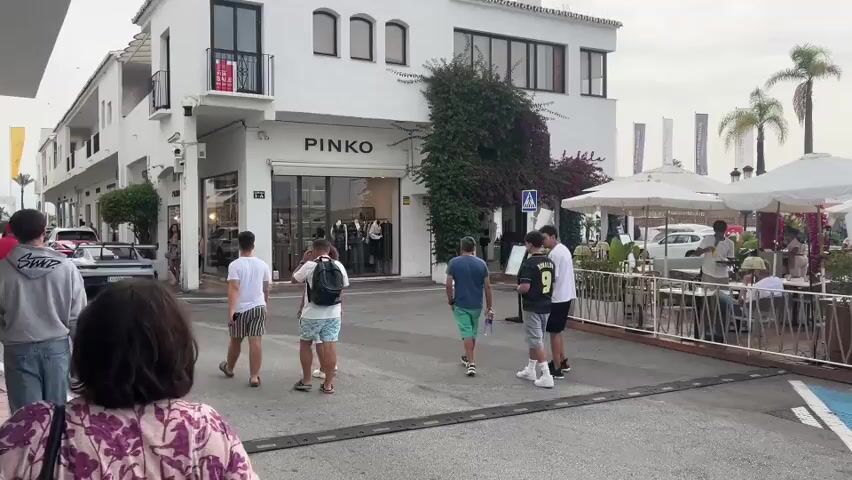 The video shows a waterfront scene with several people walking and standing. On the left, a white building has "PINKO" signage above a storefront. Vines grow up the side of the building. Several people are walking on the pavement in front of the store and towards the right side of the frame. To the right, there are outdoor seating areas with umbrellas and tables, suggesting a restaurant or cafe. A signpost with multiple directional signs is visible. In the background, there is a marina with numerous boats docked. A man in blue shorts and a white t-shirt is standing near a bin on the right side of the frame. Another man in a blue shirt is in the foreground on the left. The sky is overcast.